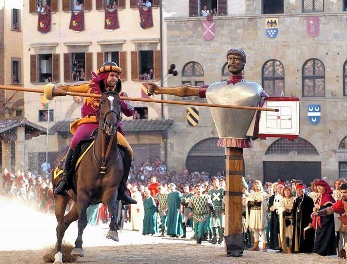 En mand på en hest i galop med et spyd i hånden, udenfor banen står en række publikummer klædt i middelalderligt tøj. 