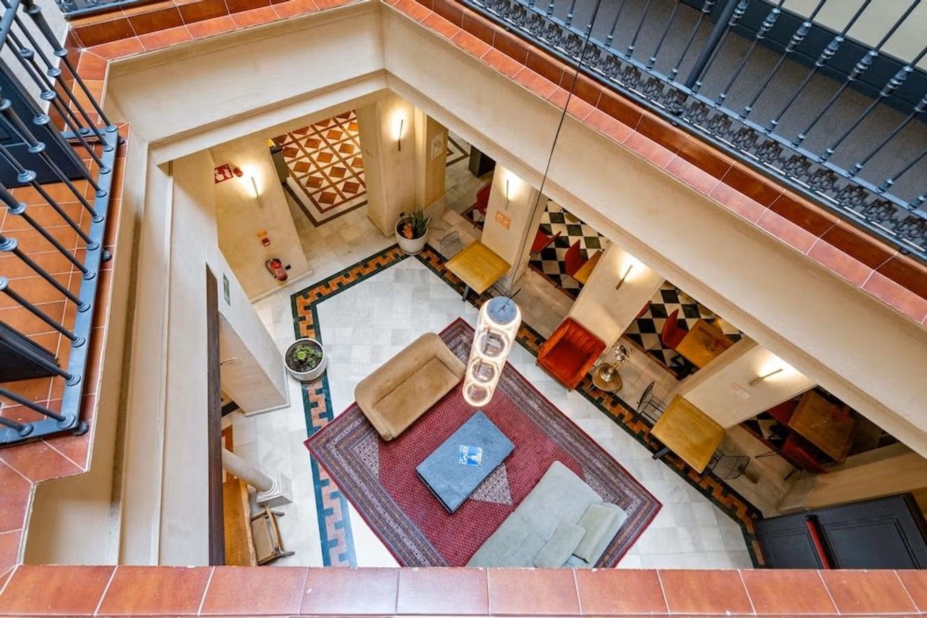 Bildet viser det åpne fellesarealet på Joya Del Casco Boutique Hotel sett ovenifra. Nederst ser vi en sofagruppe på rødt teppe, flislagte gulv og smijerns-rekkverk mellom etasjene.