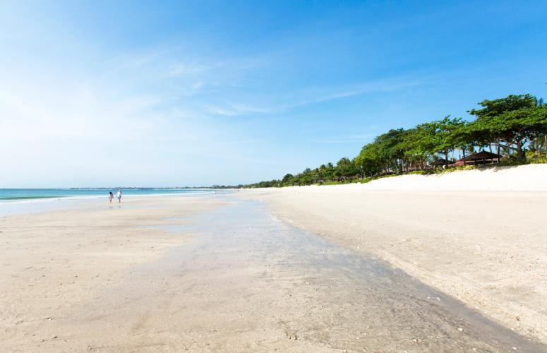 To mennesker går på stranden ved Jimbaran, mens bølgerne ruller ind. Træer, hoteller og hytter ligger langs kysten under en blå himmel.