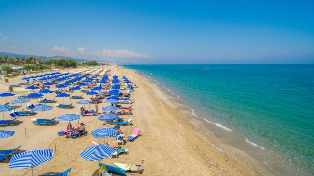 Bildet viser en flere kilometer lang sandstrand, Platanias Beach, hvor vi nærmest ser en rekke mennesker som slapper av på utplasserte solsenger med parasoller. Til høyre ligger den stille sjøen med en skyfri blå himmel over seg.