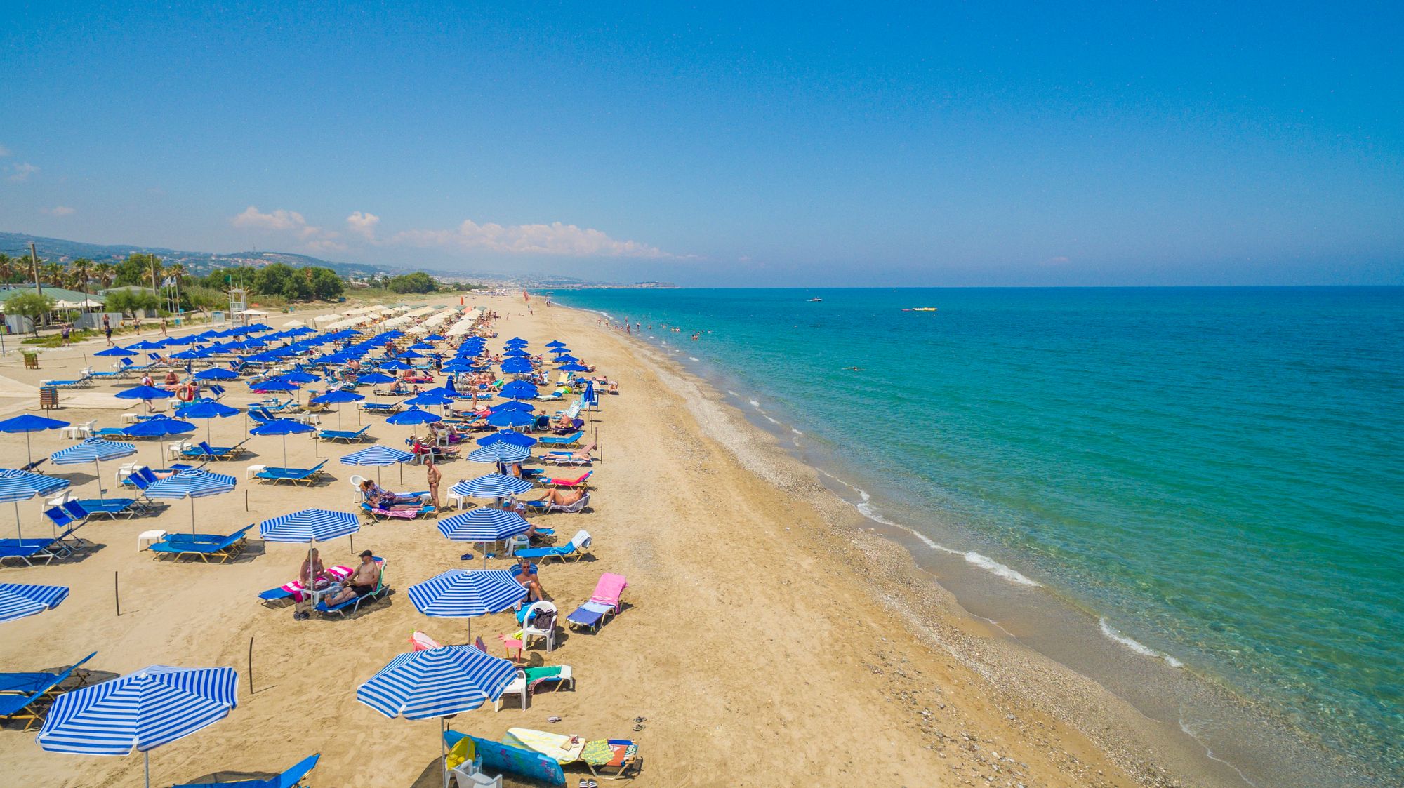 Bildet viser en flere kilometer lang sandstrand, Platanias Beach, hvor vi nærmest ser en rekke mennesker som slapper av på utplasserte solsenger med parasoller. Til høyre ligger den stille sjøen med en skyfri blå himmel over seg.