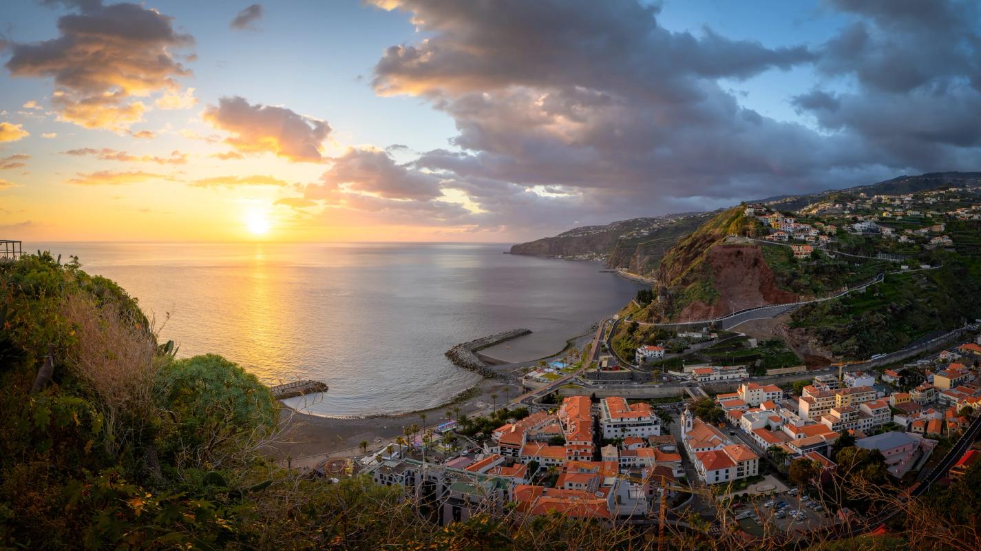 Solnedgång över bukten vid Ribeira Brava. Stadens hus har röda tegeltak och in mot staden ligger två små stränder. Staden är omgiven av klippor och i bakgrunden syns mer bebyggelse. Ett lågt molntäcke hänger över landskapet, medan havet badar i solljus.