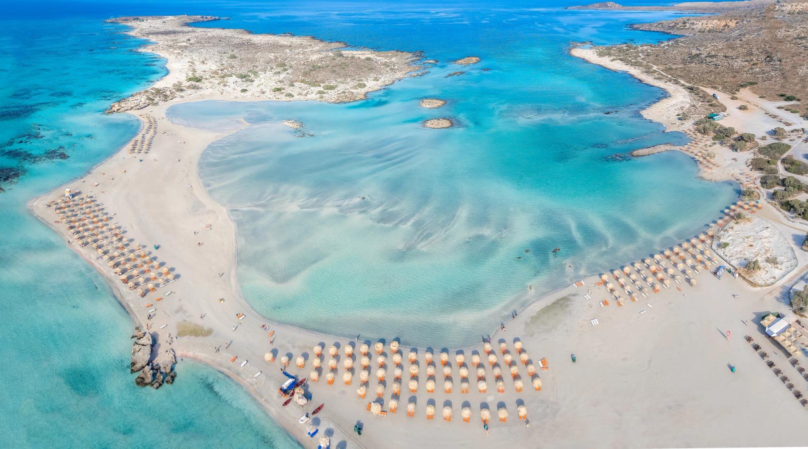 Panoramautsikt over Elafonissi Beach på vestkysten av Kreta med turkisblått vann og rosa sanddyner danner en naturlig bukt med parasoller og solsenger, mens vannet er blikkstille og krystallklart. 