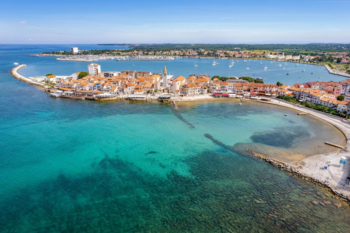 Gamla stan i Umag ligger vid det klara turkosblå havet, med tätt byggda hus med röda tegeltak, små båtar i bukten och en bred strandpromenad längs kusten.