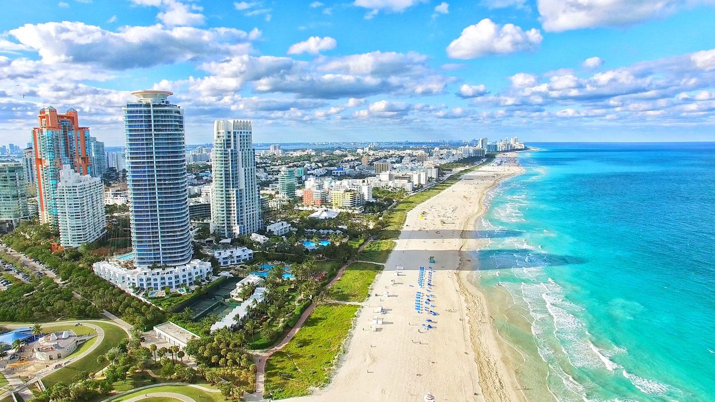 Turkosblått hav och vit sandstrand vid Miami Beach. I bakgrunden syns höga skyskrapor omgivna av gröna träd och små parker. Längs horisonten skymtar flera byggnader som följer kustlinjen.