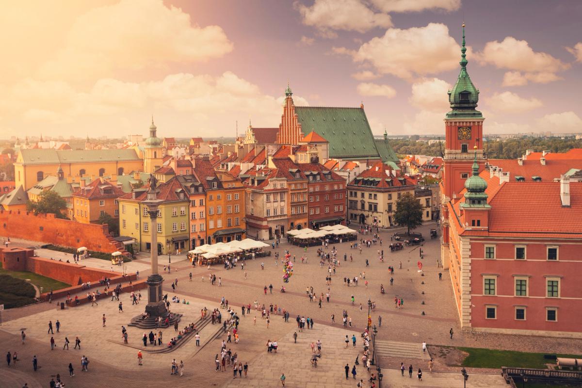Bildet viser utsikt over slottsplassen i gamlebyen i Warszawa. Midt i bildet den åpne plassen med mennesker gående. Rundt plassen eldre bygningsmasse. Himmel med spredte skyer.