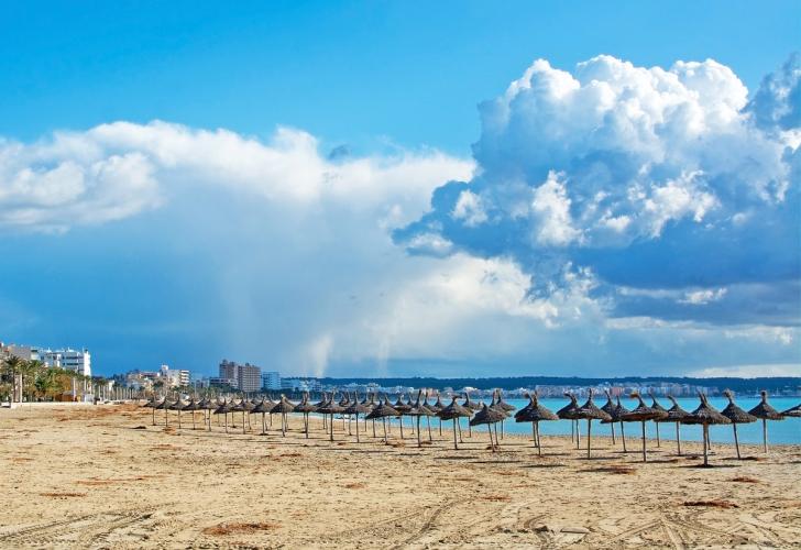 Bildet viser stranden Playa de Palma med faste parasoller i sanden. Stranden strekker seg langsetter bukten med bebyggelse i bakgrunnen og Can Pastilla lengre i enden av bildet.
