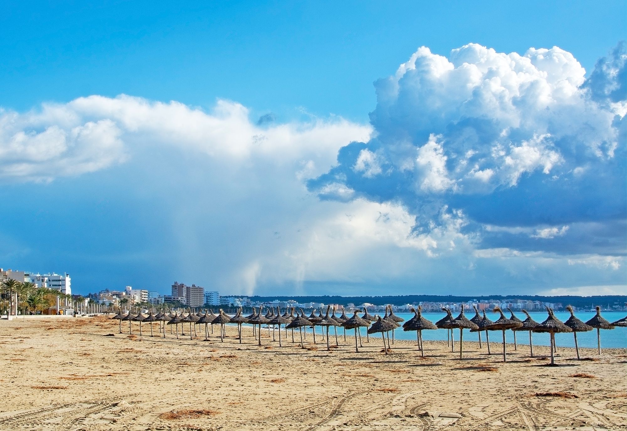 Bildet viser stranden Playa de Palma med faste parasoller i sanden. Stranden strekker seg langsetter bukten med bebyggelse i bakgrunnen og Can Pastilla lengre i enden av bildet.