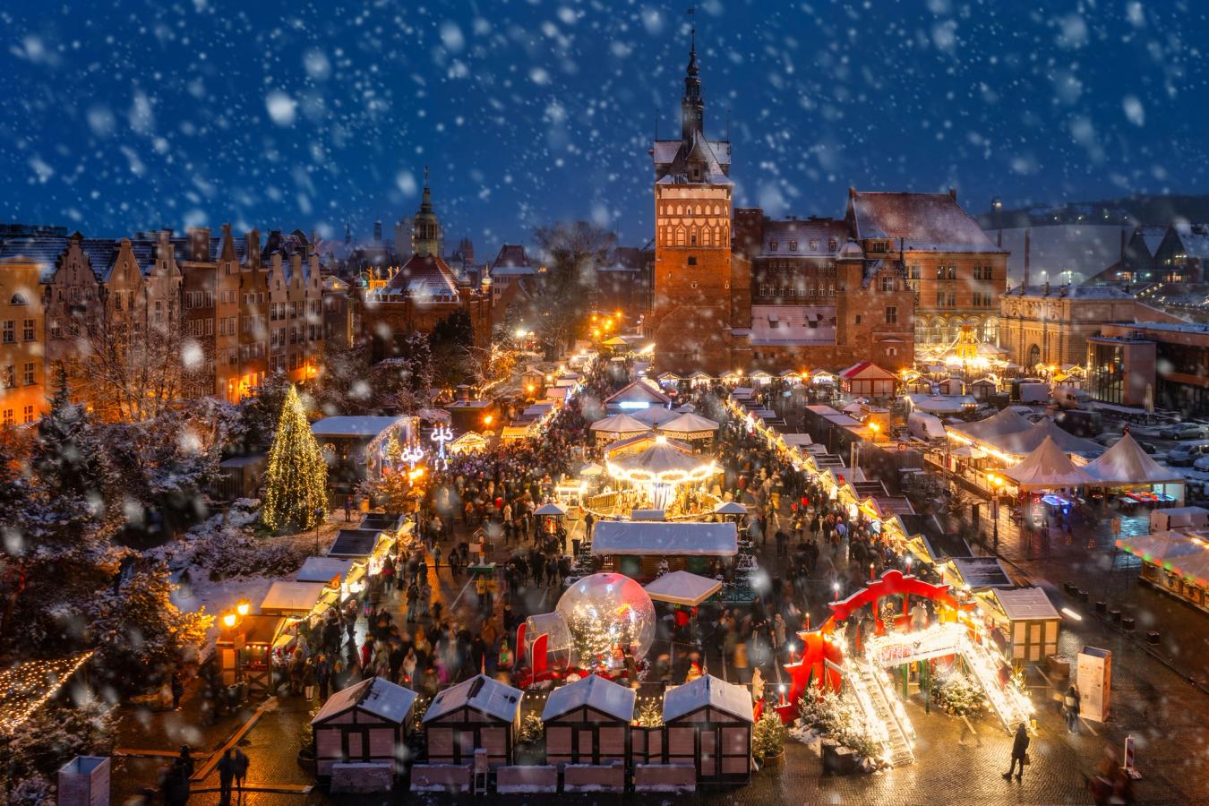 Bildet viser julemarkedet i Gdansk sett fra høyden. Det er kveld, mørkt og all julebelysning er på, med parallelle gater av salgsboder. I midten av markedet er det mindre karuseller og lett snødrev i lufta.