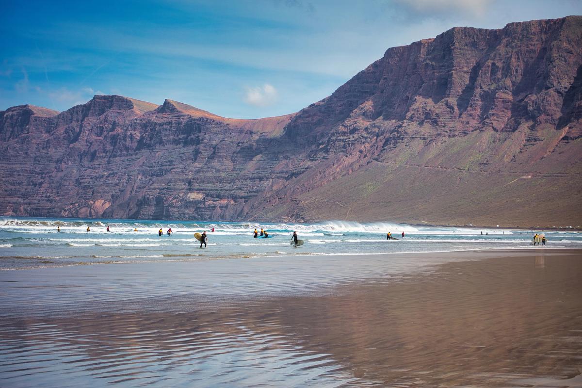 Billedet viser stranden Playa de Famara med flere aktive surfere og kitesurfere på vej ind og ud af havet. Stranden er omgivet af klippeformationer.