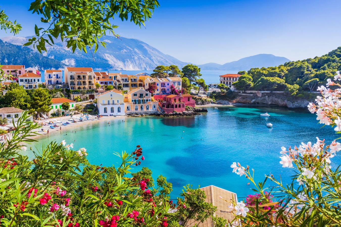 Bilden visar den lilla byn Assos på ön Kefalonia. Man kan se små en- och tvåvåningshus i olika färger. Det kristallklara vattnet liknar en lagun mot byggnaderna, där stranden är central i stadsbilden. De små byggnaderna är omgivna av blommor och frodig natur, en kristallklar himmel och längst bort på bilden kan man se bergformationer