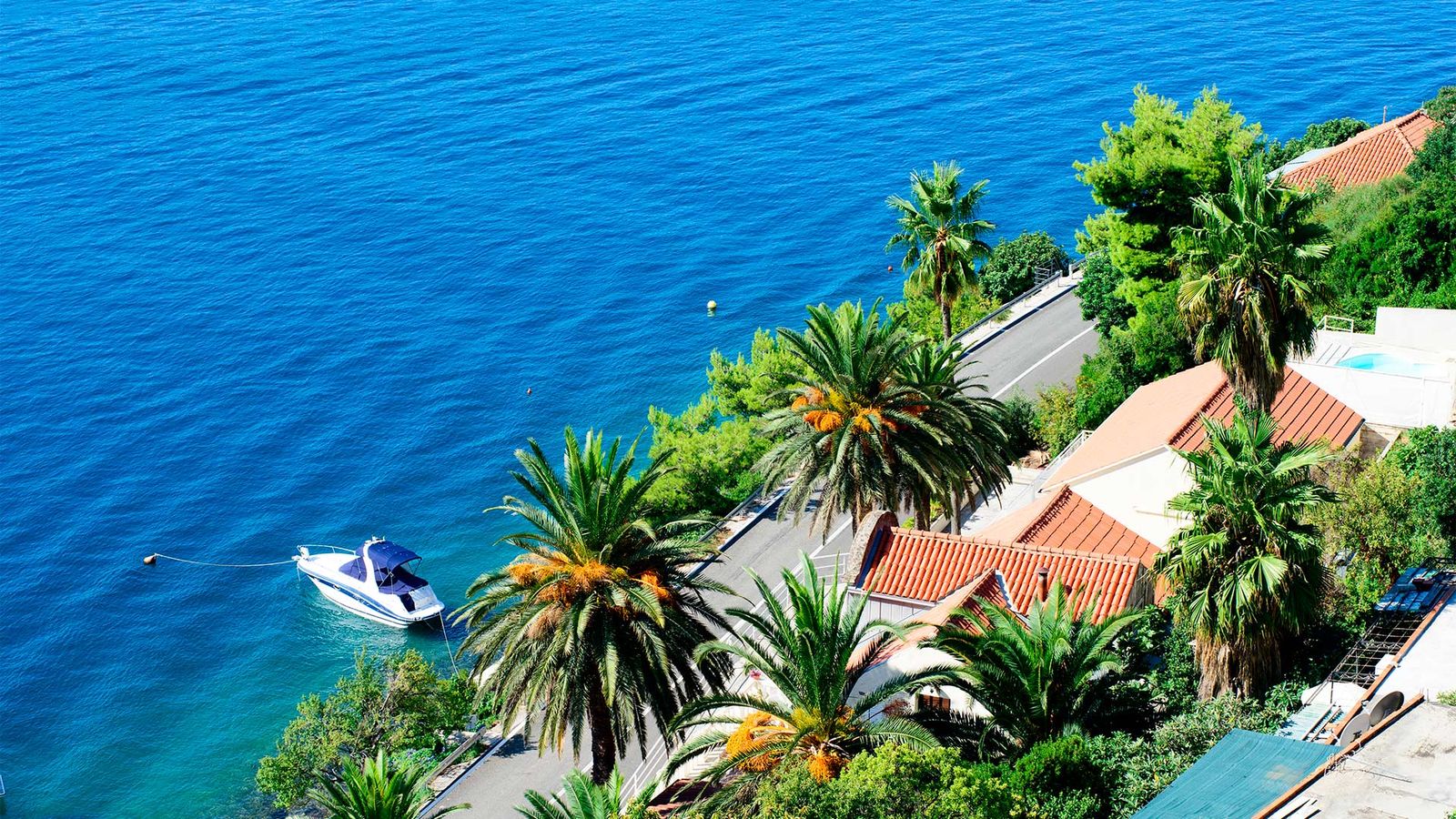 En lille båd ligger for anker i krystalblåt vand. En kystvej snor sig langs havet og langs den er der palmer og anden vegetation samt et hvidt hus. 