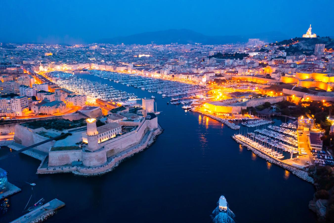 Billedet viser den gamle havn i Marseille om aftenen. Indgangen til havnen har en fæstning til venstre og en større marina indenfor, fyldt med både. Bag havnen ser vi hele bybilledet i aftenlys.