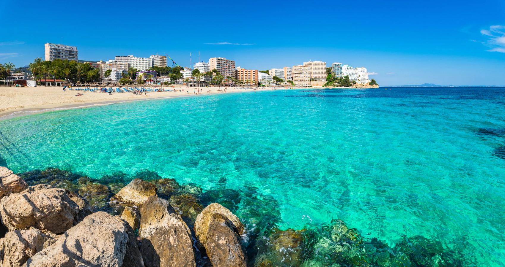 Bilden visar huvudstranden Platja de Magaluf sedd från havet. Först syns några klippor i vattnet, sedan längre in i bilden ser man det lugna vattnet som slutar i stranden med parasoller och badgäster. Längst bak i bilden ser vi byggnader från staden Magaluf. Allt under en blå och molnfri himmel