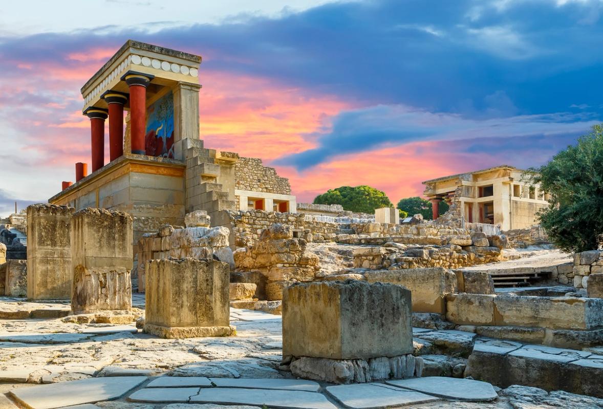 Bildet viser ruinene etter Knossos Palace ved byen Heraklion. På bakken er det gamle steinheller med rester av bærekontruksjoner rundt om. Midt i bildet vises det mest ivaretatte av ruinene, en gjenstående vegg med bæresøyler i rød farge. Himmelen er på kveldstid med en rødlig solnedgang.