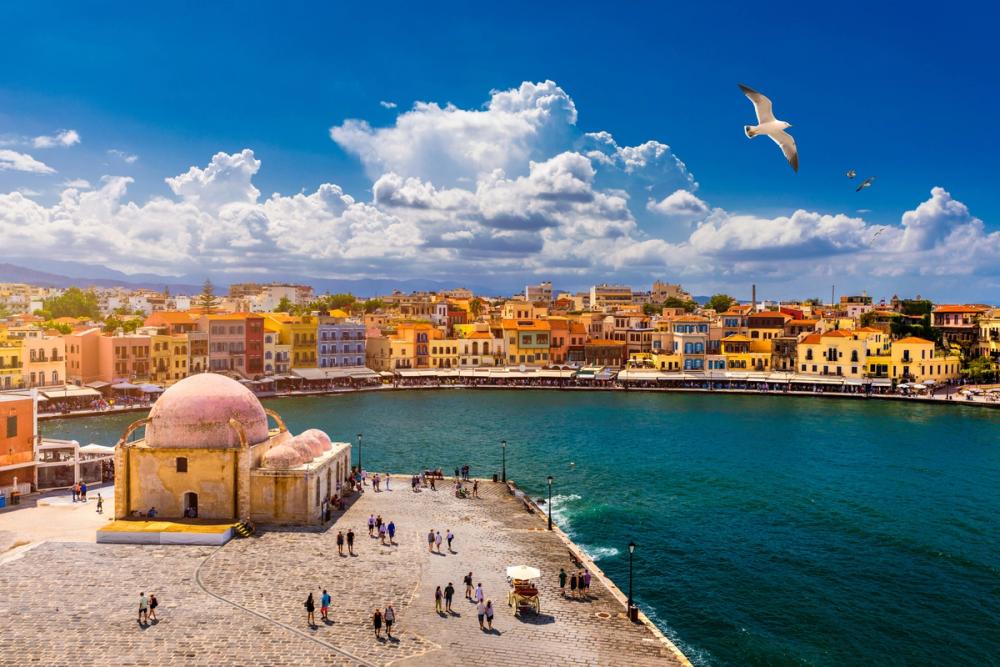 Bildet viser Den Venetianske havna i byen Chania på Kreta. Fremst i bildet ser vi den mindre tyrkiske moskeen på hjørnet av havnebebyggelsen. Videre innover i bildet ser vi strandpromenaden som er omgitt av en lav okergul bebyggelse. Øverst i bildet flyr en måke med blå himmel som har enkelte spredte skyer.