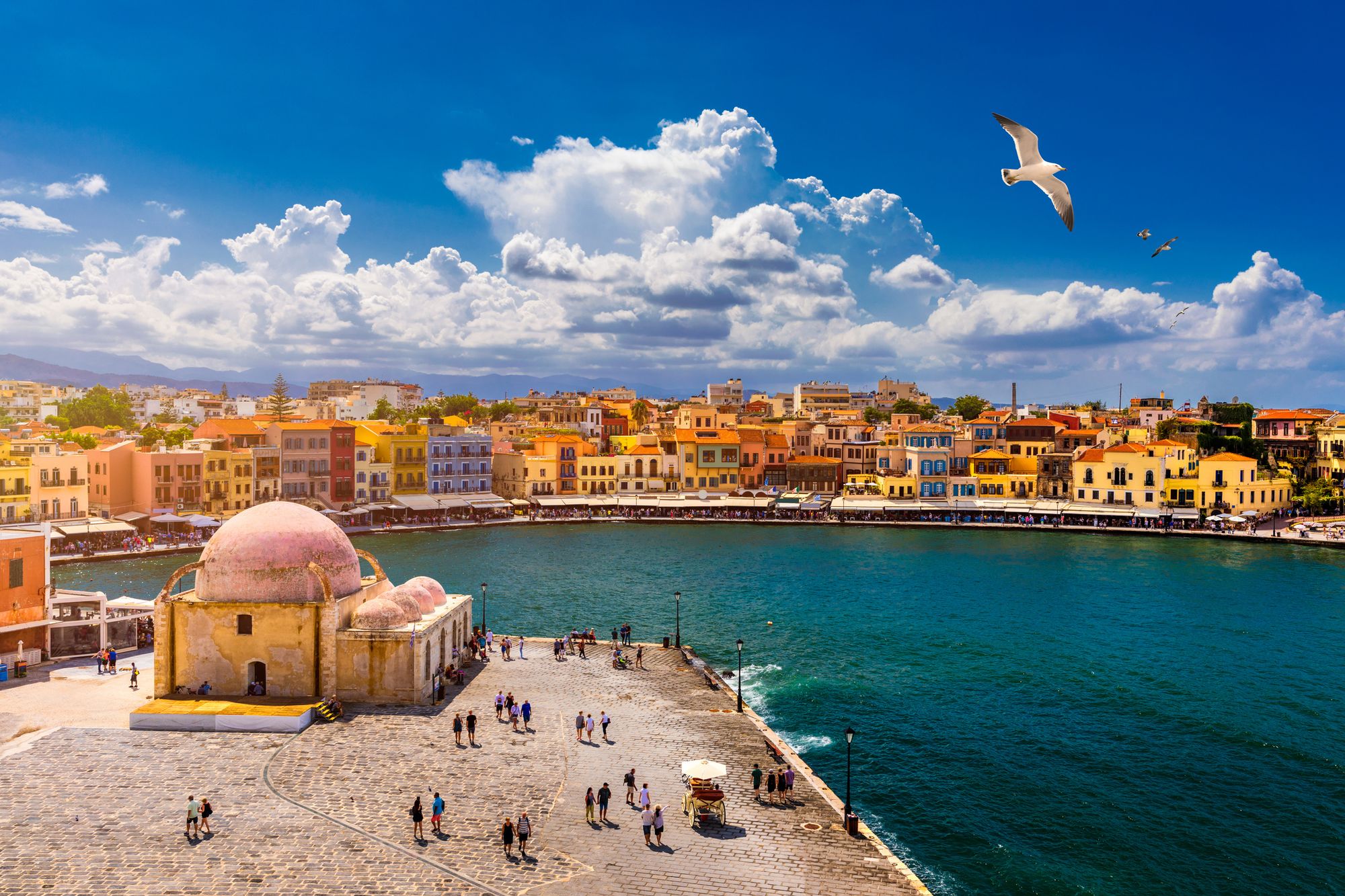 Bildet viser Den Venetianske havna i byen Chania på Kreta. Fremst i bildet ser vi den mindre tyrkiske moskeen på hjørnet av havnebebyggelsen. Videre innover i bildet ser vi strandpromenaden som er omgitt av en lav okergul bebyggelse. Øverst i bildet flyr en måke med blå himmel som har enkelte spredte skyer.