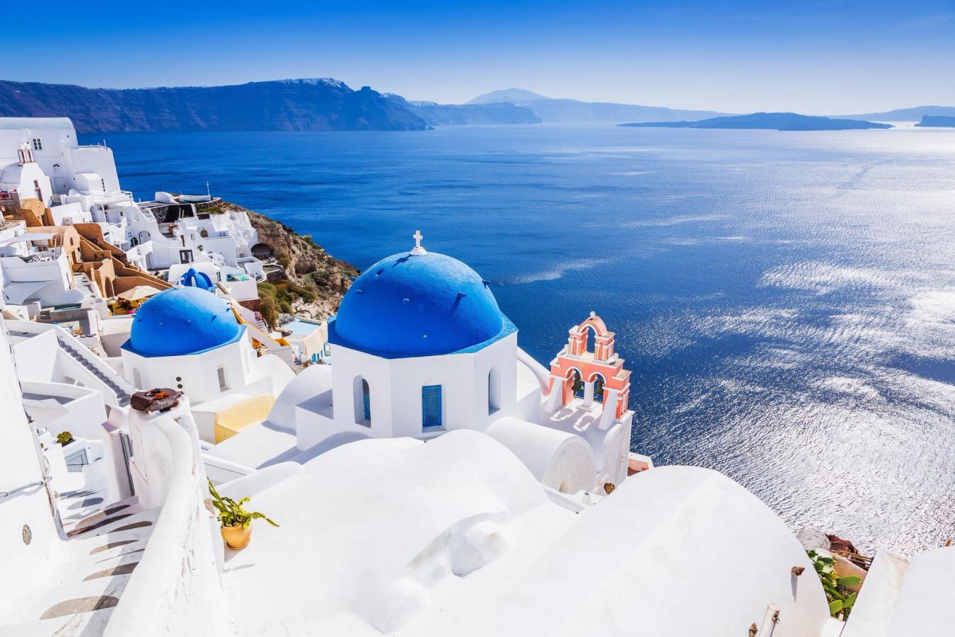 Fotot är taget på ön Santorini i Grekland och visar de klassiska vitkalkade husen längs klippsluttningen med sina koboltblåa tak och kupoler. Byggnaderna slutar vid ett lugnt, blått hav med en molnfri himmel