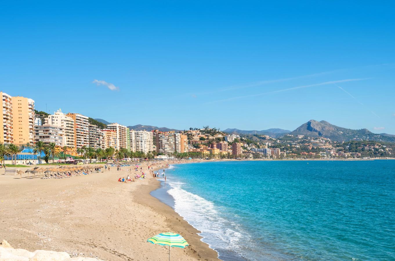 Bildet viser den langgrunne stranden Playa de La Malagueta, som er bystranda i Malaga. Bakenfor stranda, til venstre, ser man bybebyggelse og lengst inn i bildet vises fjellformasjoner.