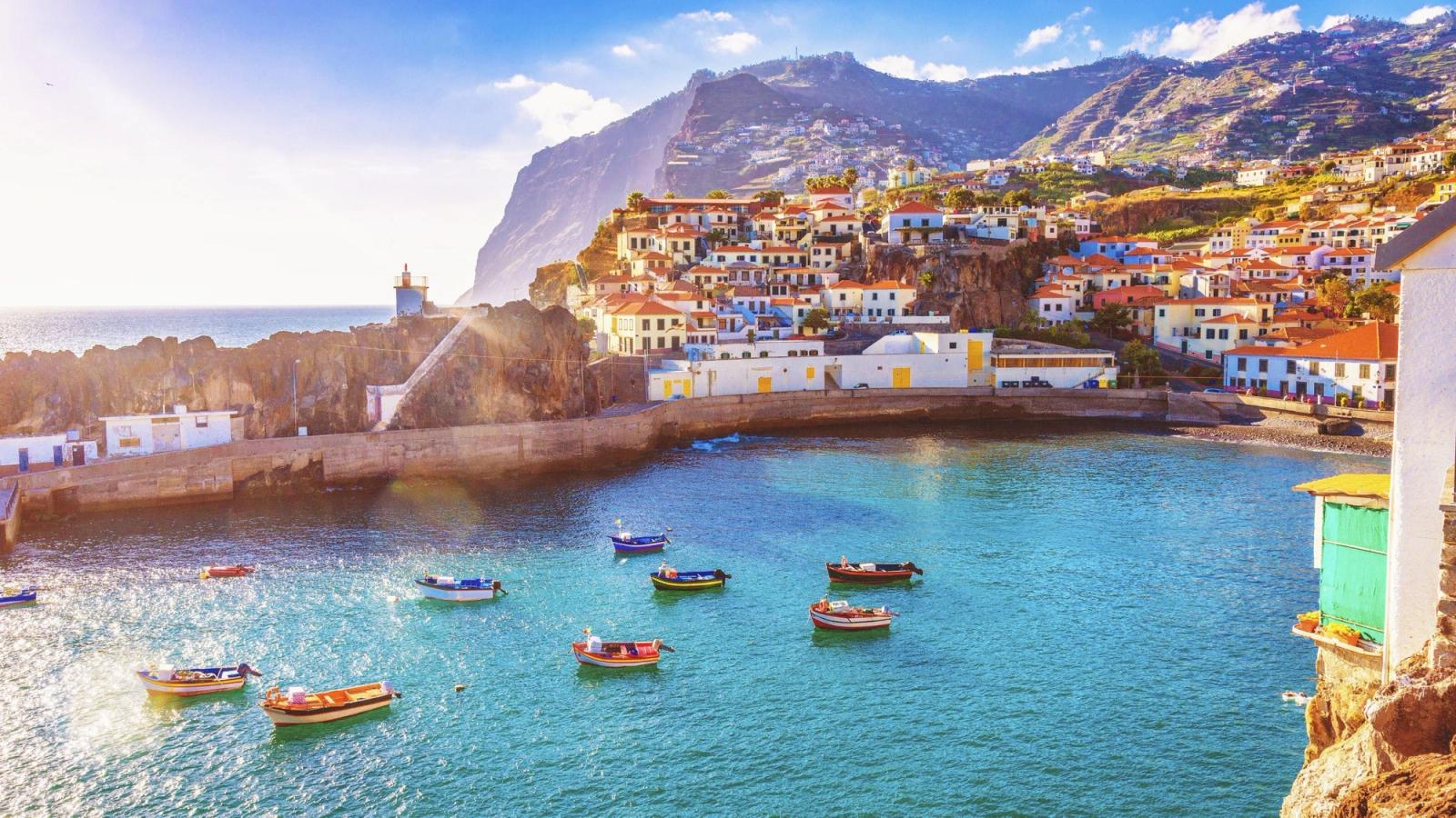 I en lille bugt med turkisblåt vand ligger flere fiskerbåde i forskellige farver fortøjet. Flere hvidkalkede huse med røde tegltage strækker sig op langs bjergskråningerne. I baggrunden rejser stejle klippeskråninger sig mod den blå himmel. 