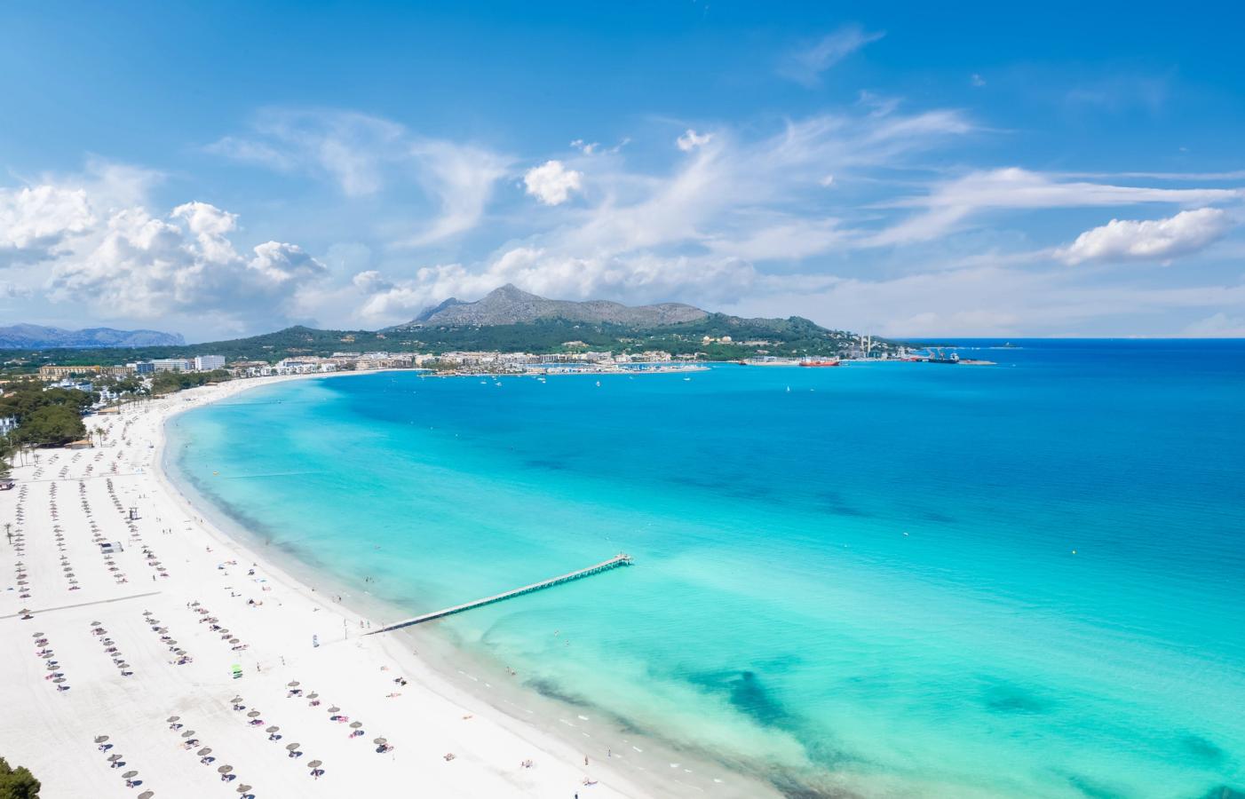 Billedet viser stranden Playa de Muro i Alcudia-bugten, lige nord for Can Picafort. Vi kan se det krystalblå Middelhav mod den næsten hvide sandstrand, der strækker sig flere kilometer ind i billedet. Yderst ser vi klipperne ved "enden" af Mallorca på nordsiden. Alt sammen under en blå himmel med spredte skyer.