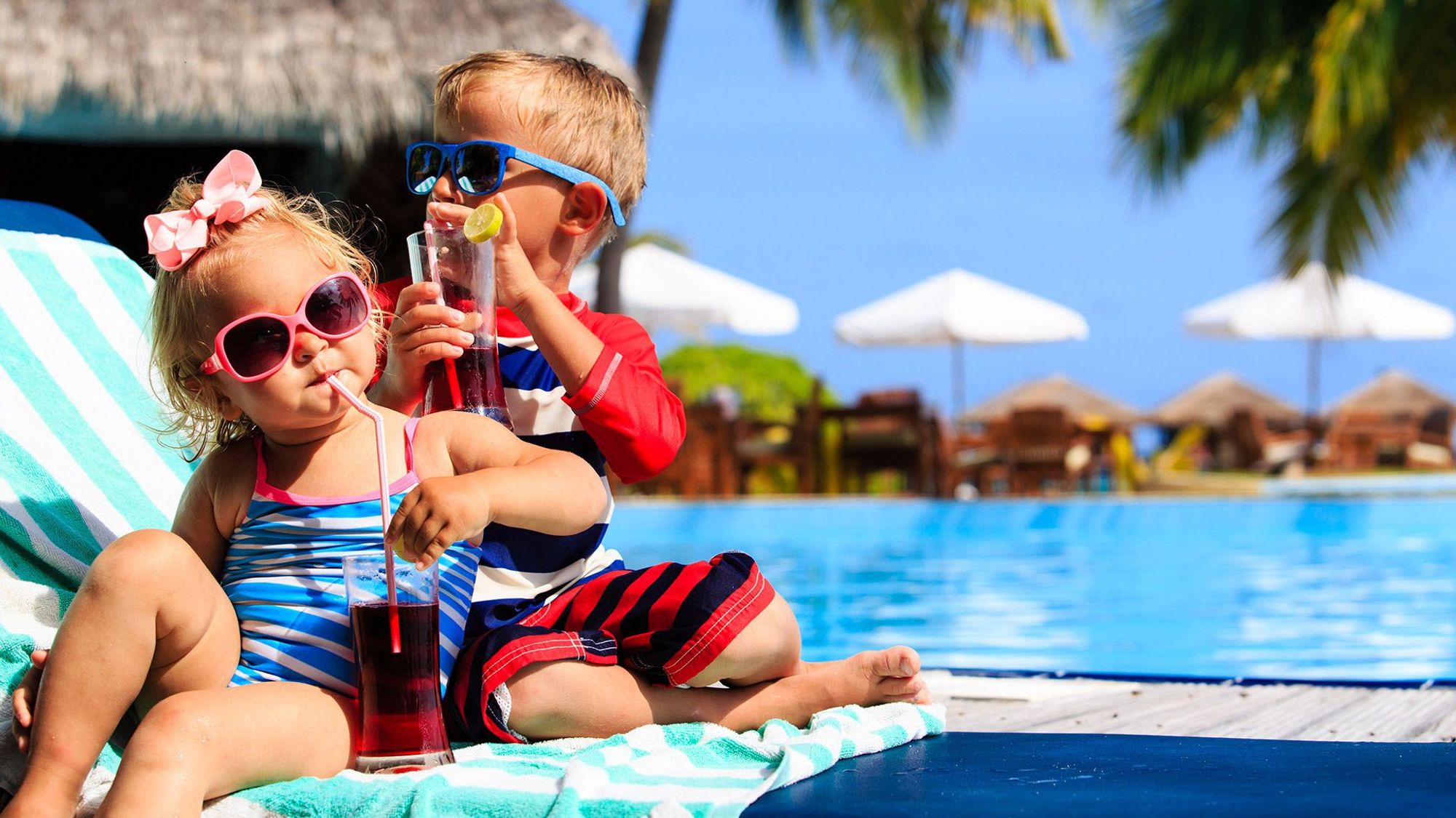 Bildet er av to små barn sittende på en solseng med hvert sitt glass med drikke og sugerør. I bakgrunnen et svømmebasseng.