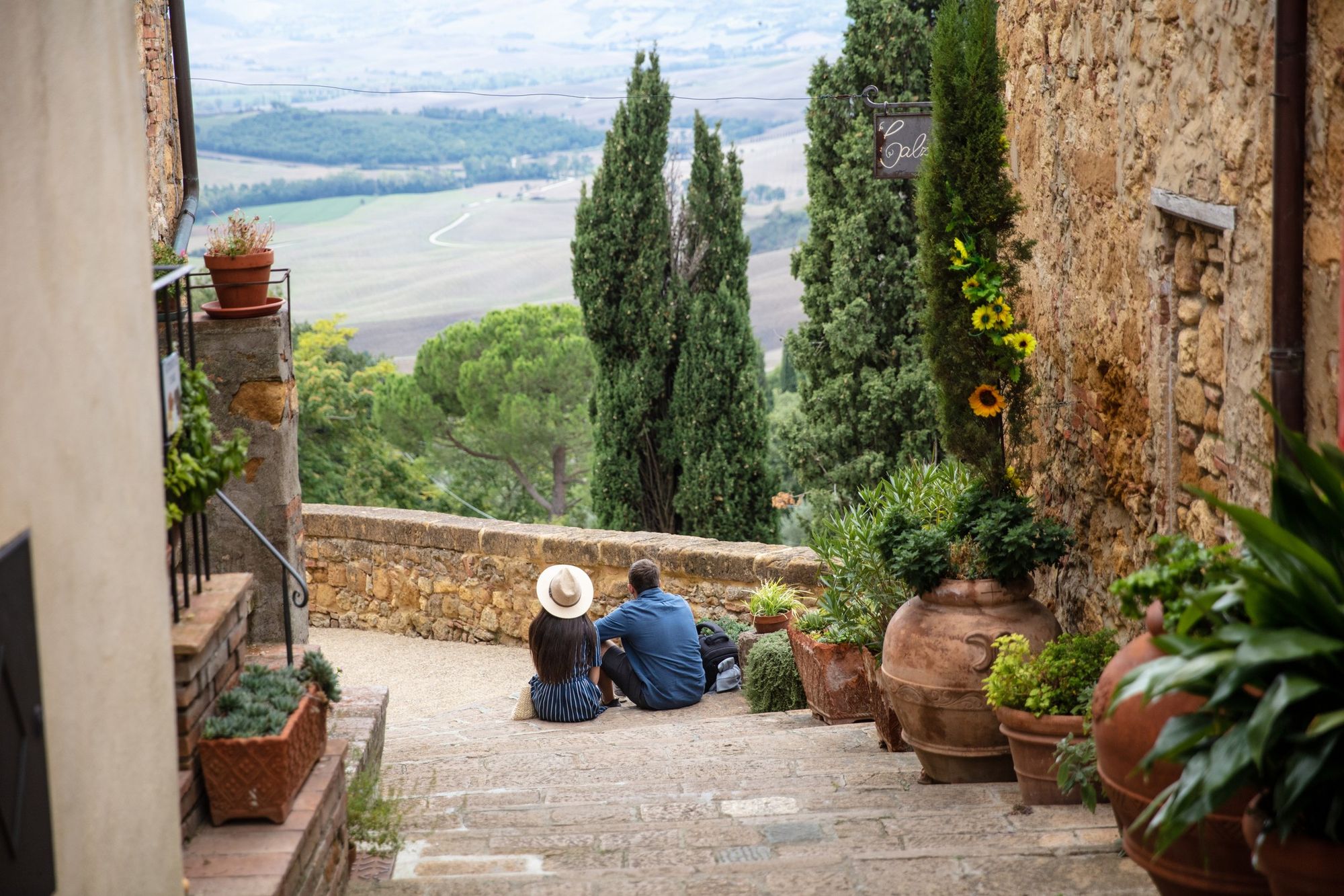 Et par, en mand og en kvinde, sidder på en trappe mellem ældre beboelsesejendomme og nyder udsigten over store sletter i Italien. Frodig vegetation og beplantning langs husmurene.