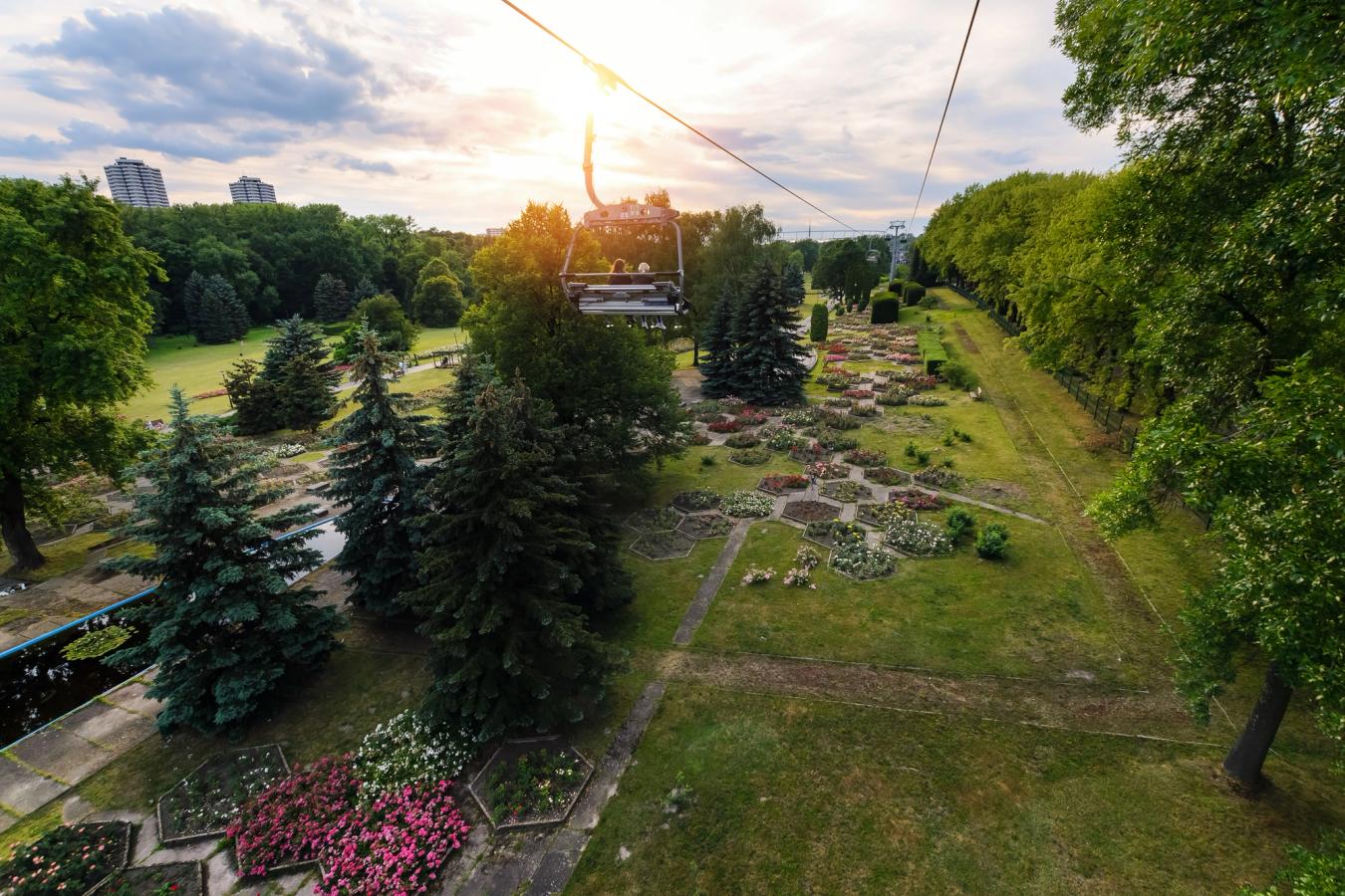 Billedet viser en svævebane med gondolvogne, der kører gennem Silesian Park i Katowice. Vognene hænger lavt over parken, hvor vi kan se en vandoverflade og flere farverige blomsterbede på jorden. På begge sider af svævebanen findes parkanlæg og frodige træer.