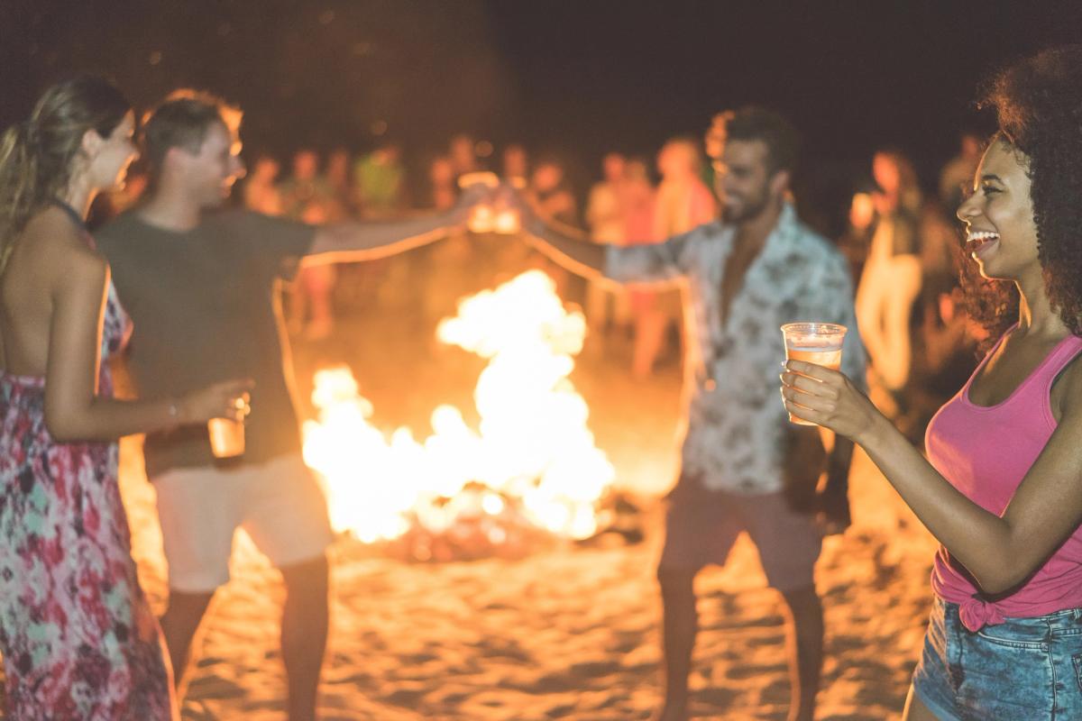 Bilden visar unga människor som festar på en strand på natten. I mitten av festglada ungdomar brinner en bål.