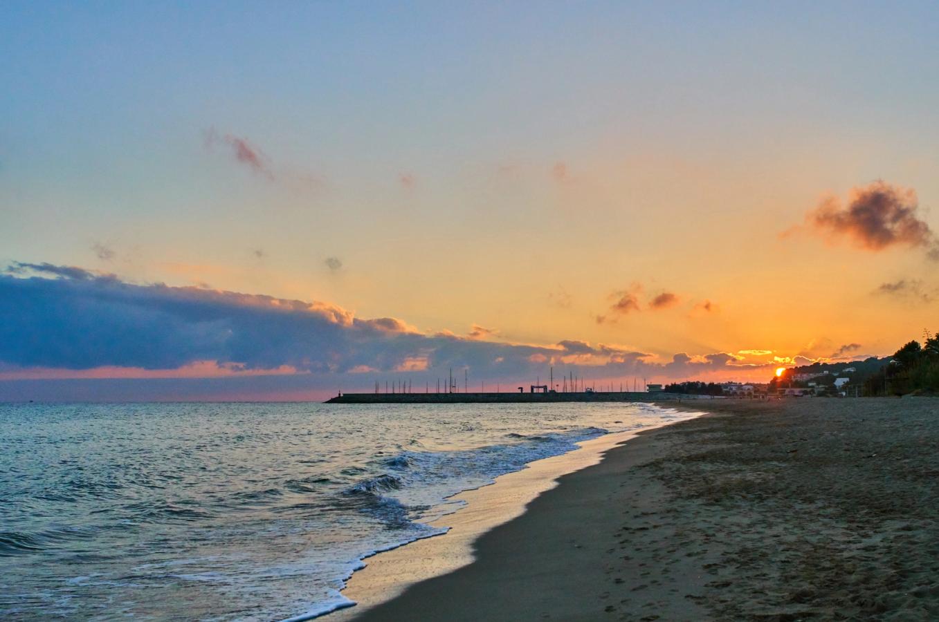 Solnedgang ved stranden i Coma-Ruga med rolige bølger, mørkt sand og himmel i varme farver.