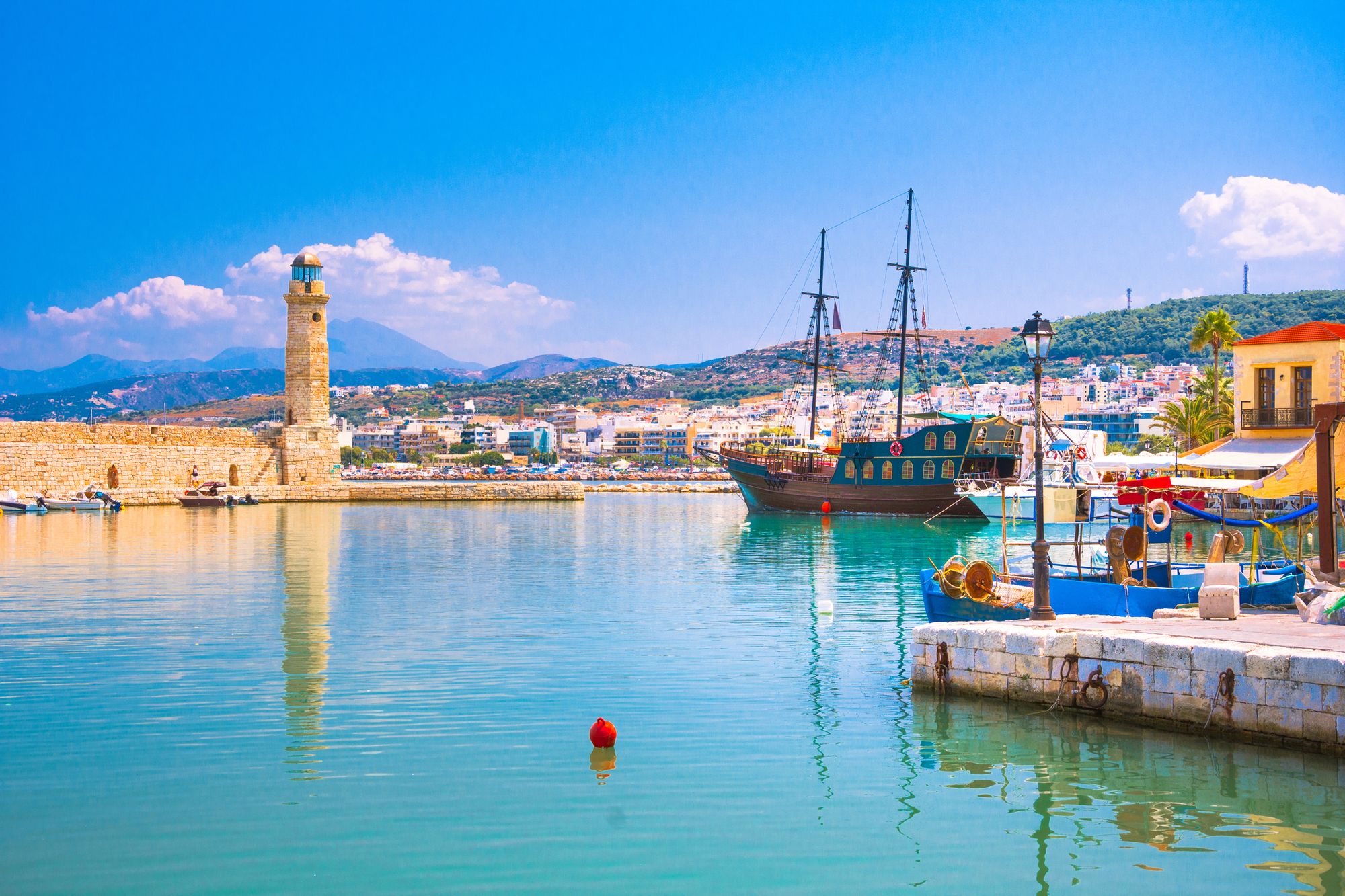 Bildet viser den gamle venetiske havna i Rethymnon på Kreta. Ført i bildet, nederst til høyre, ser vi en liten utstikker med en liten kafe og en blå fiskebåt fortøyd. Lenger ut i vannet ligger en kopi av en gammel venetiansk seilskute med to master. På venstre side ser vi en større del av den eldre havna med et fyrtårn. Lengst inn i bildet ser vi bebyggelsen til byen Rethymnon på Kreta. Himmelen er blå med små spredte skyer.