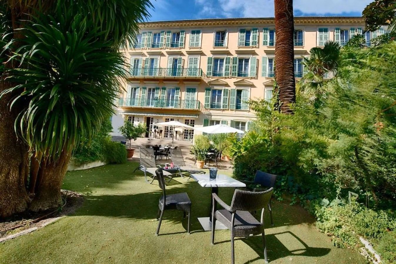 Bildet viser hotellets hage, fra baksiden av fasaden. I en frodig hage med kaffebord, palmer, parasoller. Midt i bildet hotellets bakside med balkonger og grønnmalte vinduskarmer.