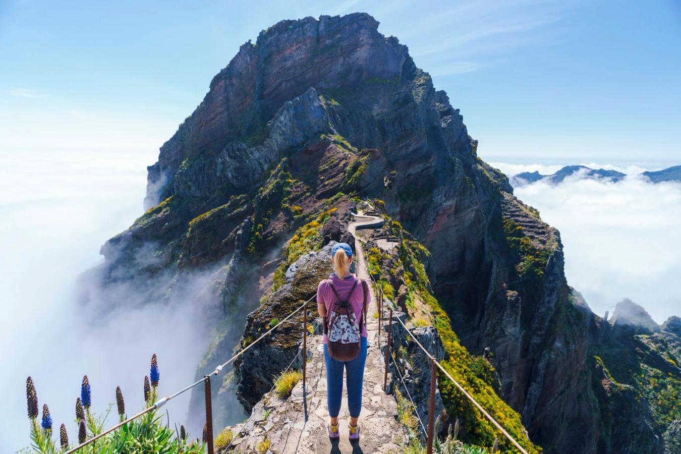 En kvinne er på vei mot fjelltoppen Pico do Arieiro på Madeira. Turstien er tilrettelagt med tauverk på hver sin side. Fjelltoppen er klippepreget og skogkledd, og ligge rover øverste skylag.