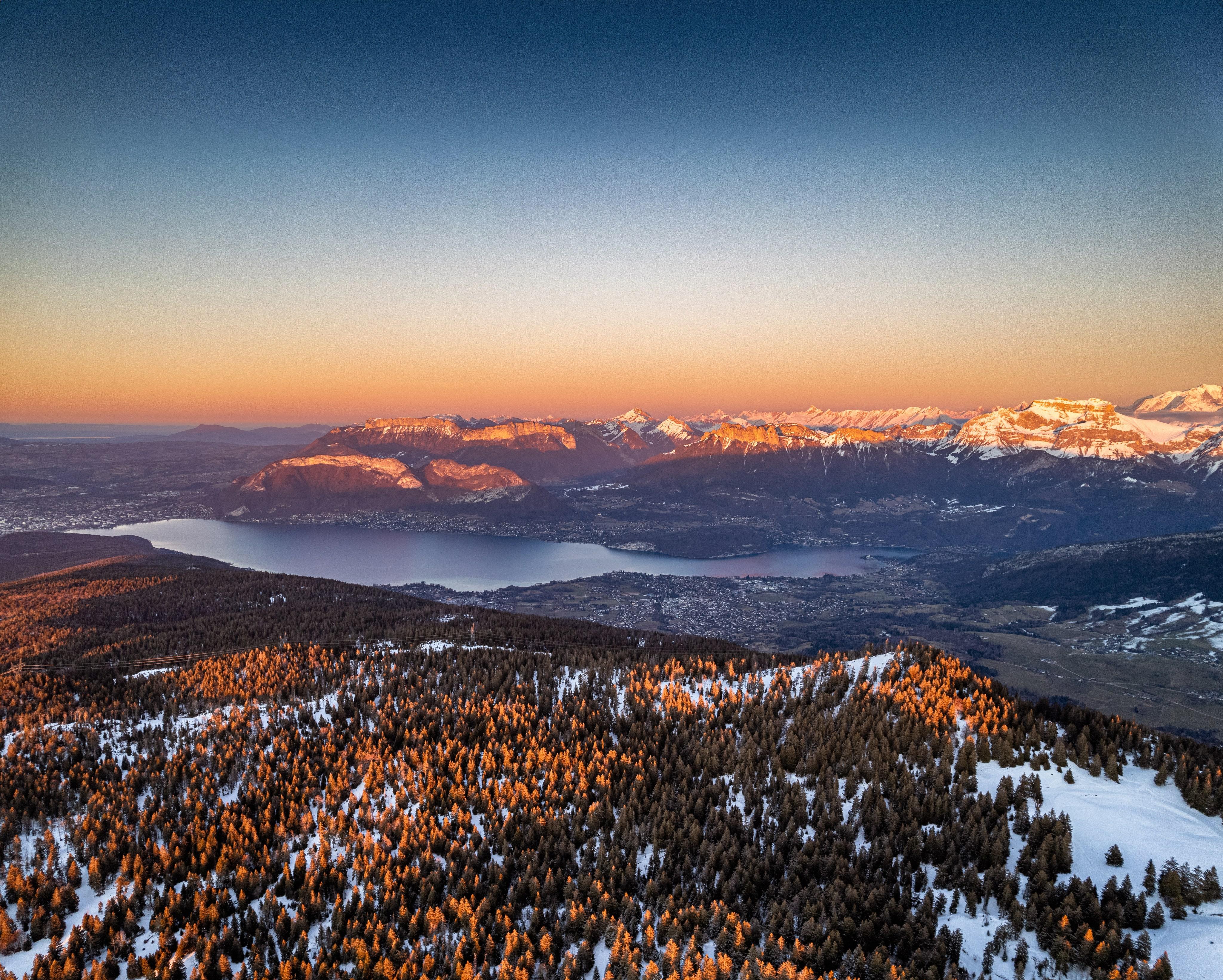 Vue aérienne au coucher de soleil sur le lac d'Annecy entouré de montagnes enneigées et de forêts de mélèzes aux teintes dorées