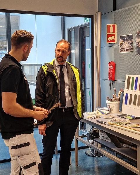 Maler i arbeidsklær i samtale med HMH Kong Haakon VIII ved fargekart, pensler og materialprøver