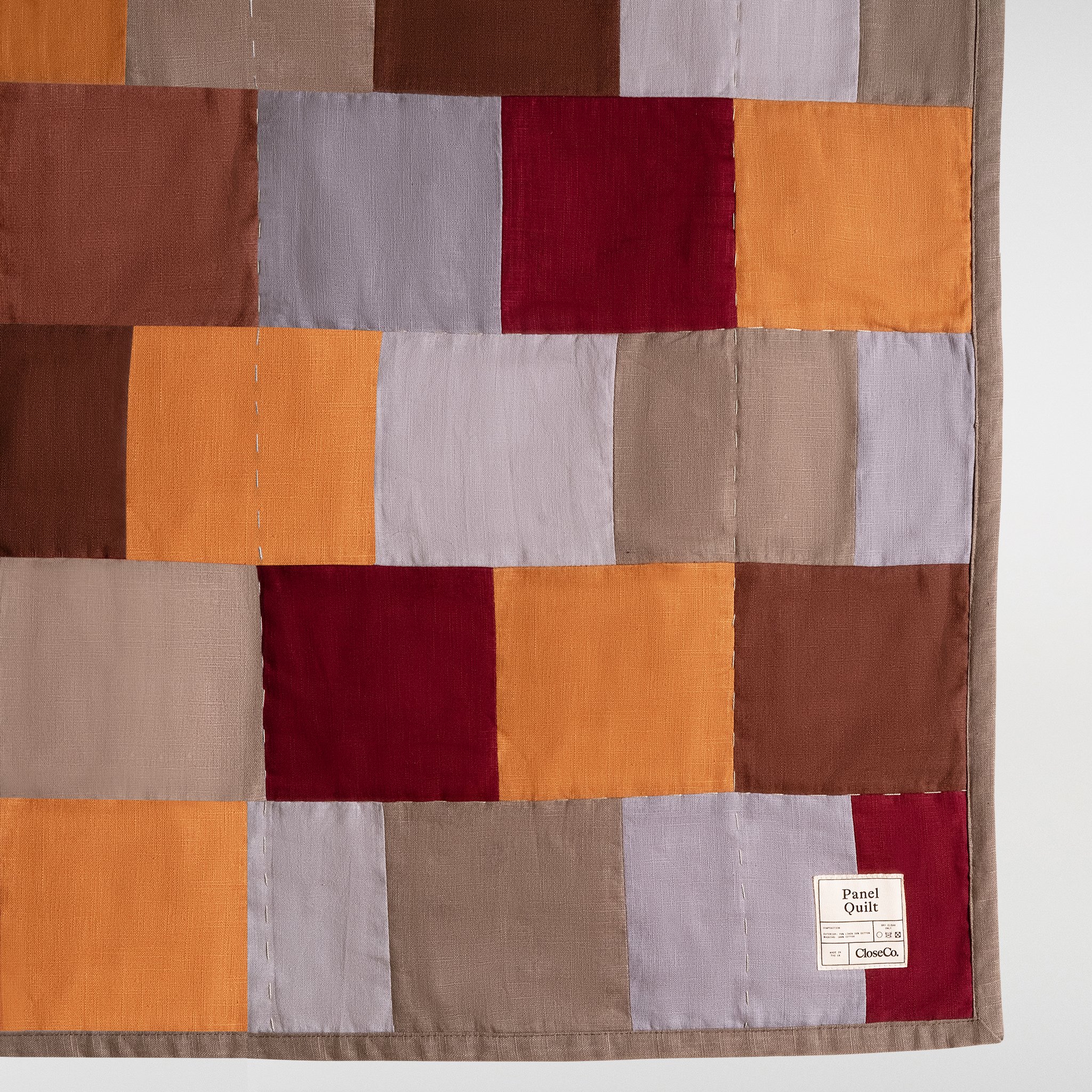 Panel patchwork quilt in warm red, rust, ochre and neutral tones, close-up detail highlighting stitched panels, fabric texture and tonal variation