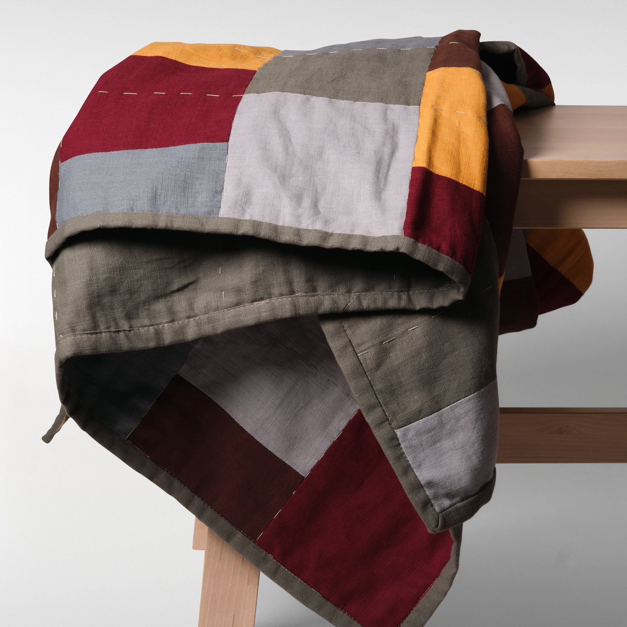 Panel patchwork quilt in warm red, rust, ochre and neutral tones, draped over a wooden bench showing soft structure, stitching and natural fabric finish