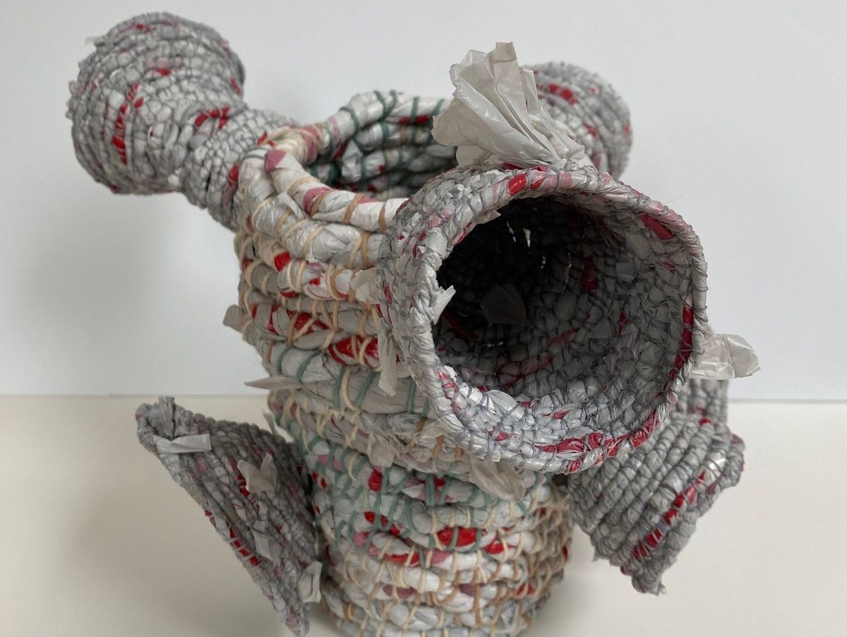 A large coiled vessel worked from glossy grey and red plastic film, with two tubular spouts branching from the body. Torn tufts of white plastic spike out between the tightly bound rounds.