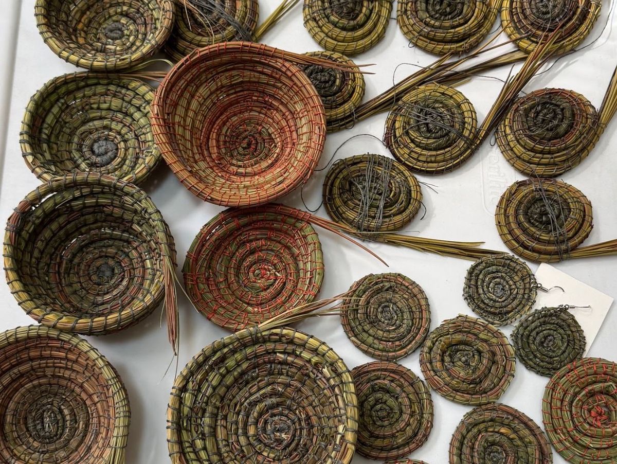 Overhead view of many coiled discs and shallow baskets laid on white, worked from flattened grasses in olive, moss and coral. Loose stem ends and dark thread trail from their edges.