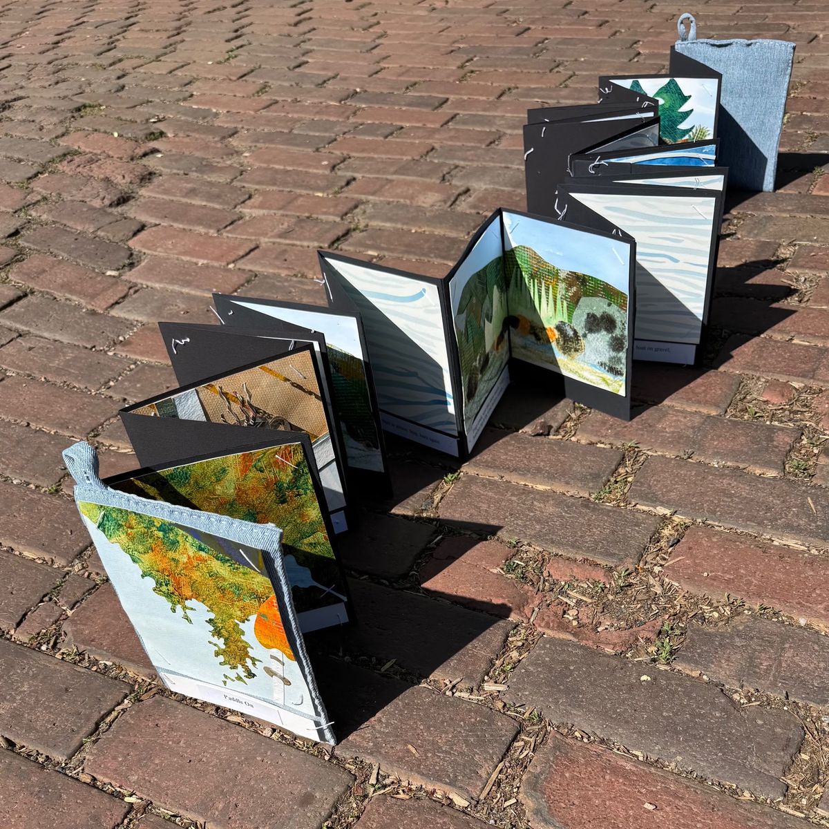 An accordion book zigzags across sunlit brick paving, casting hard shadows. Black pages frame collaged landscapes — orange and green foliage, pale water, a dark shoreline. A grey cloth slipcase stands at the far end.
