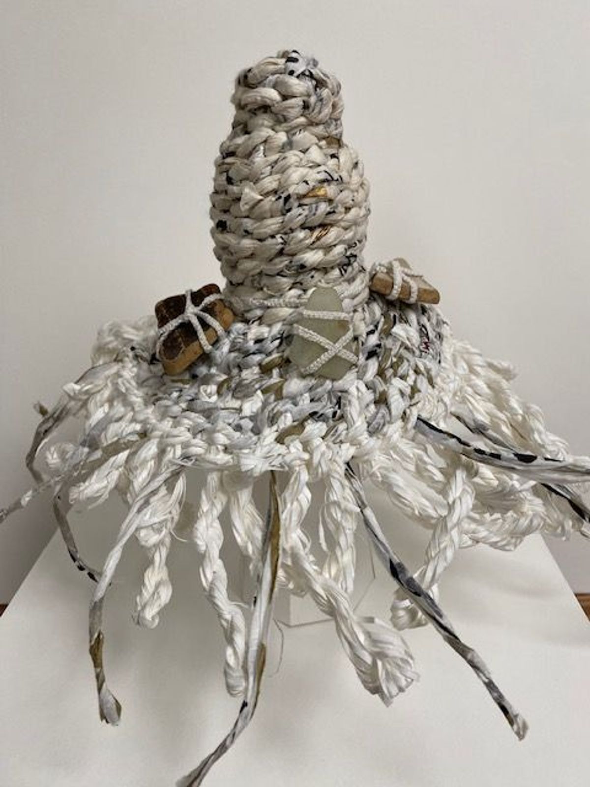 A tall conical form coiled from white paper cord, skirted with a dense fringe of twisted paper strands. Pieces of curling bark and a pale flat stone are bound to its shoulder with cord.