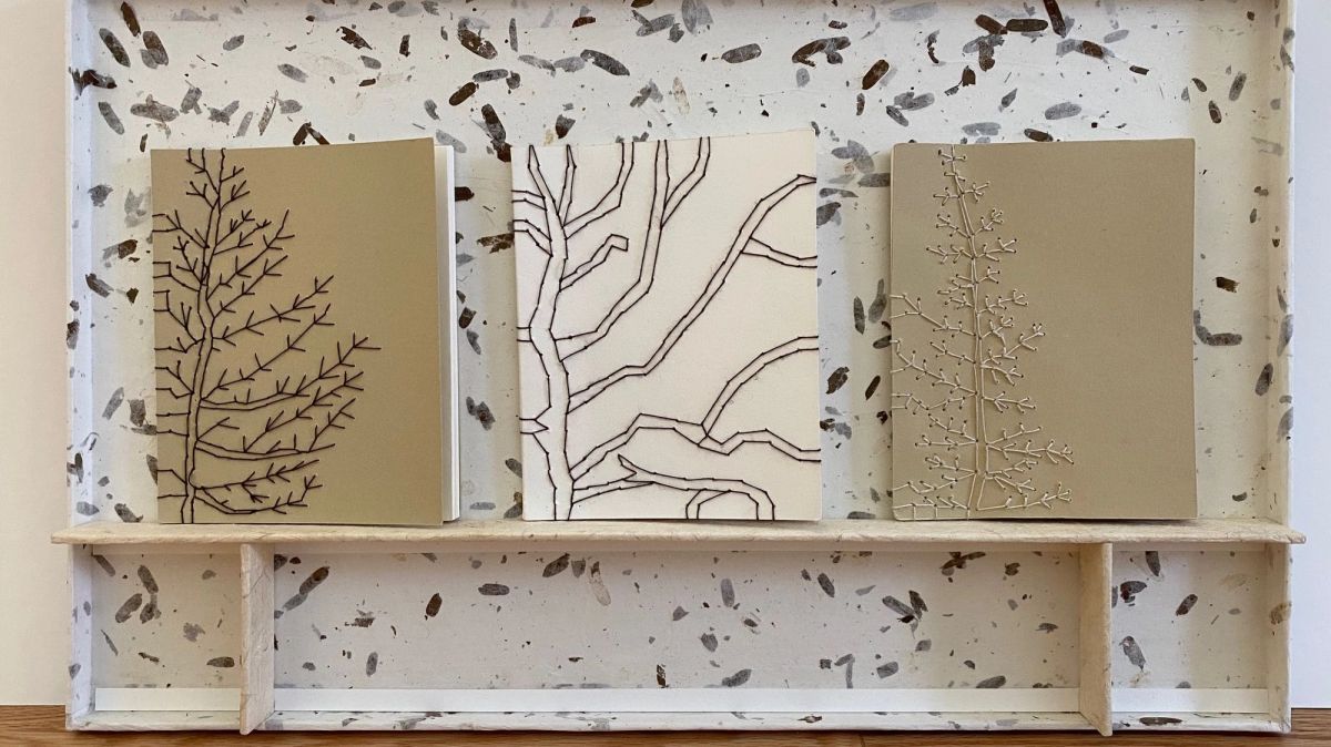A shallow white shelf box whose walls are set with pressed seed pods and leaf fragments. Three panels stand on the ledge: a brown branch drawn on olive, black branch lines on cream, and a pale raised branch in relief.