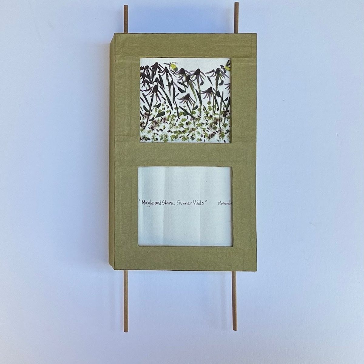 A moss-green covered panel with two cut windows, suspended between slim wooden dowels. The upper window frames a pressed botanical print of dark stems and green leaves; the lower holds a handwritten title on soft white paper.