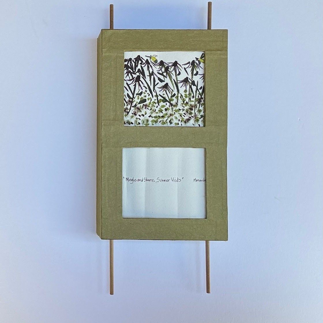 A moss-green covered panel with two cut windows, suspended between slim wooden dowels. The upper window frames a pressed botanical print of dark stems and green leaves; the lower holds a handwritten title on soft white paper.