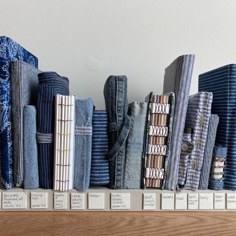 A shelf of handmade books stood upright, nearly all bound in denim and striped shirting in blues and greys. Exposed spine stitching shows on several. Handwritten tiles below label each: "Camping Trips", "Poetry", "Gardens", "Lake Poems".