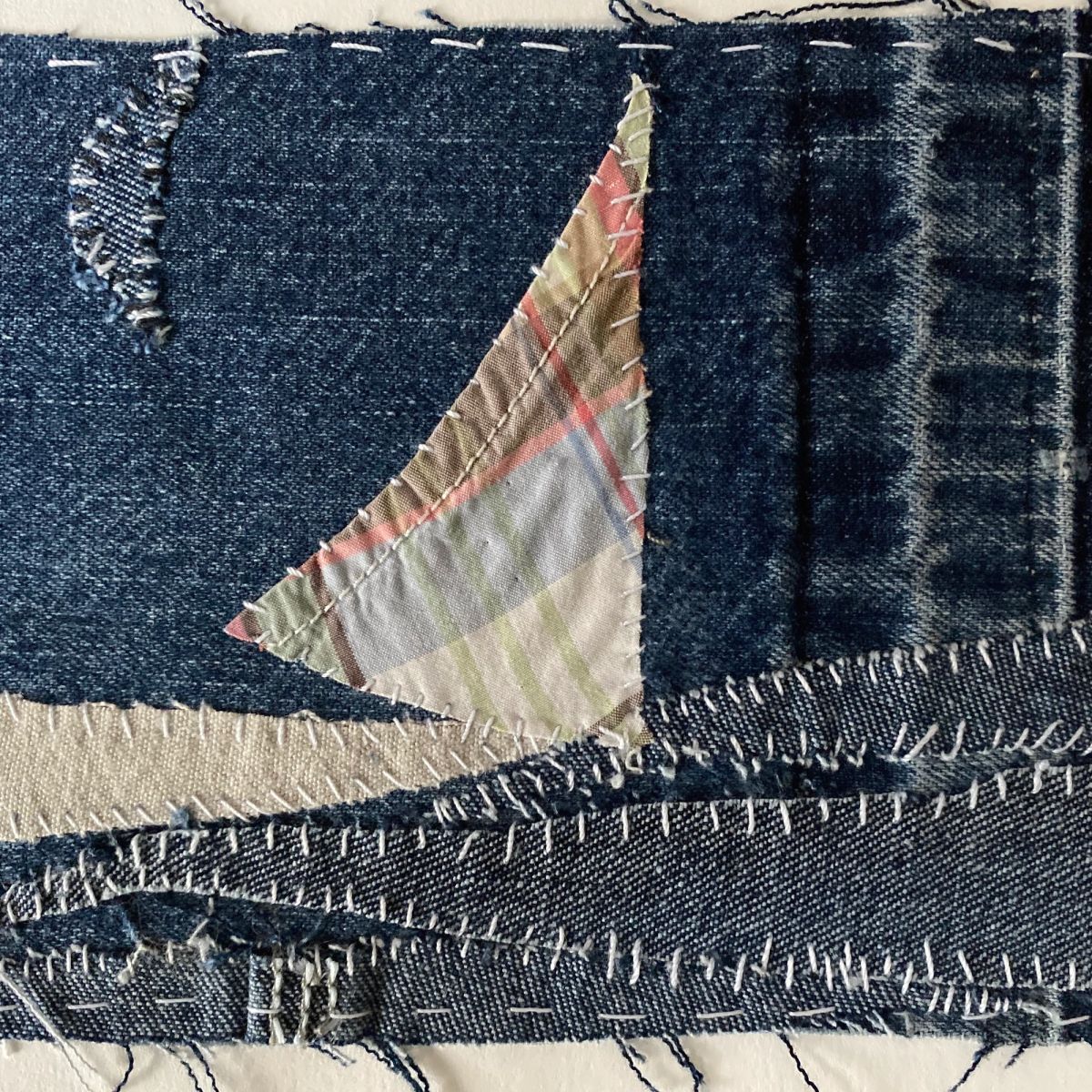 A triangular sail cut from soft plaid cotton, hand-stitched onto a ground of pieced indigo denim. Frayed denim strips layer beneath it as water, worked over with rows of white stitching.