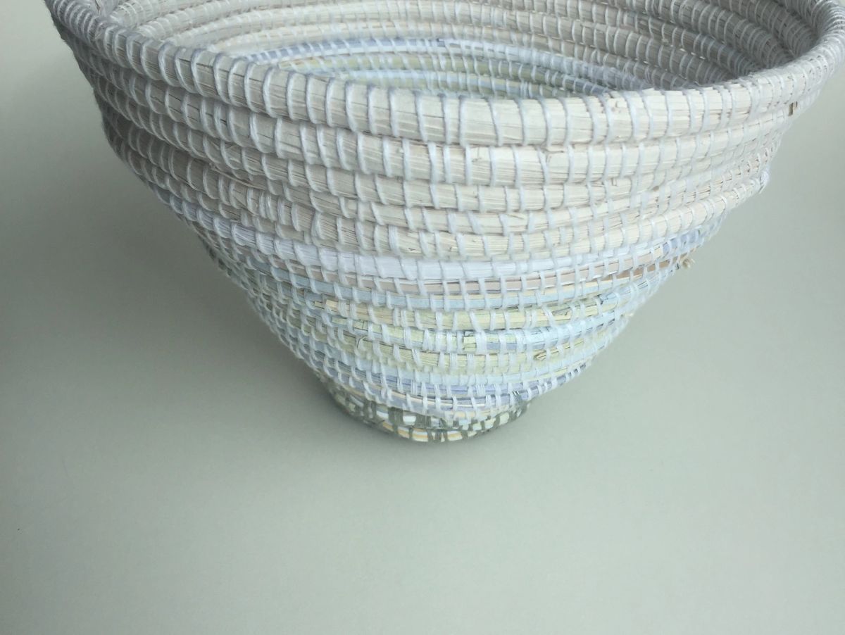 Close view of a conical coiled basket in white and palest cream, the wrapped strips stitched in tight concentric rounds. Faint flecks of printed colour surface near the base. Soft, shadowless light.