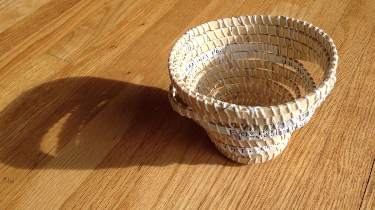A small coiled basket with a rippling asymmetric rim sits on oak in raking sunlight, casting a long soft shadow. Fragments of printed text run through the wrapped coils — "Every human", "DINER".
