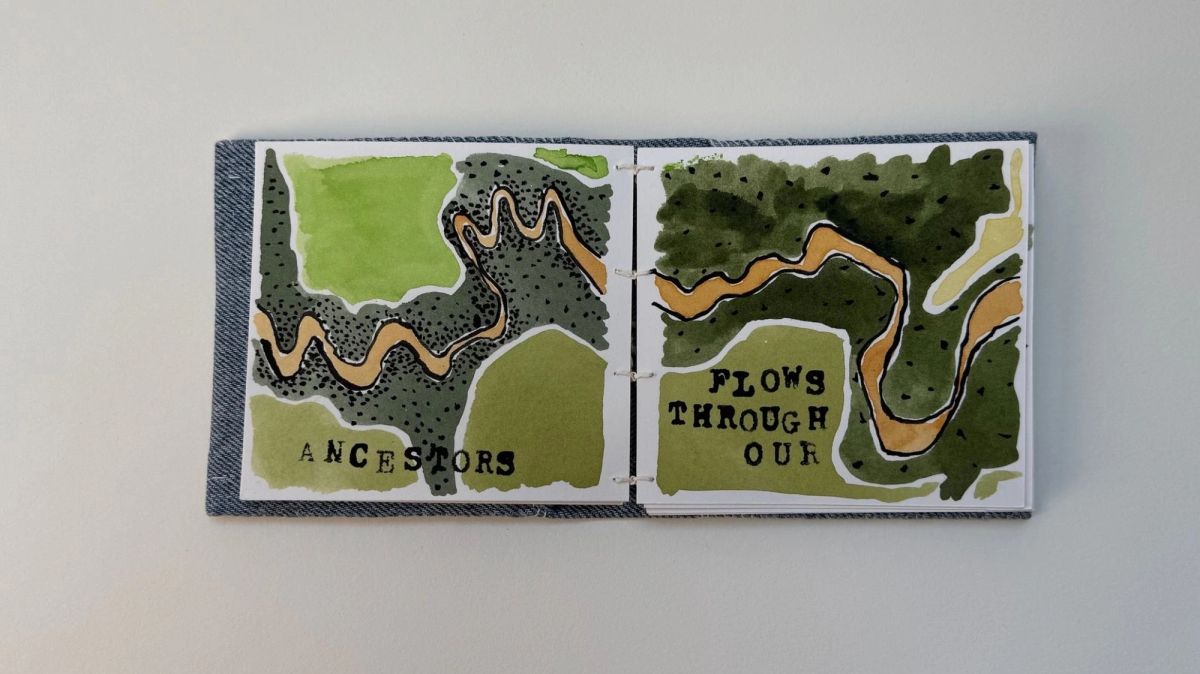 A denim-covered book open flat. Painted river maps in two greens with an ochre channel meandering across, stamped "ANCESTORS" and "FLOWS THROUGH OUR". Exposed white binding thread crosses the spine.