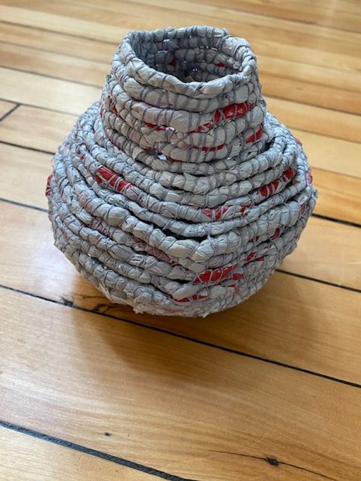 A round-bellied vessel with a narrow neck, coiled from grey printed strips shot through with flashes of red. The wrapped rounds swell outward then draw in sharply. It sits on a worn oak floor.